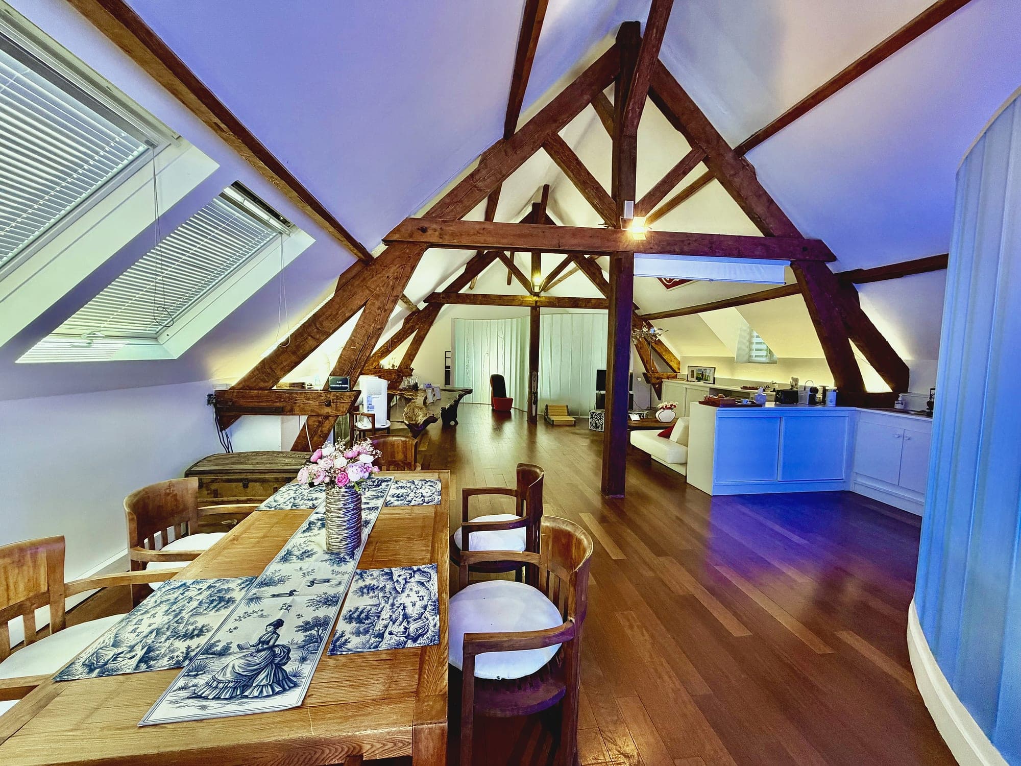Le séjour du loft sous la charpente de chêne, la table dressée et la cuisine au fond